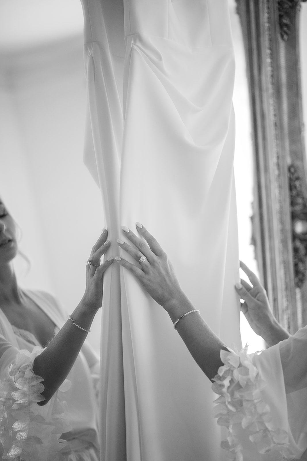 Black and white close-up of the bride's hands gently touching the fabric of her hanging wedding dress.